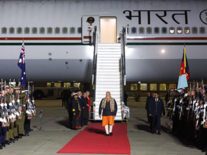 Dignitary walks a red carpet away from a large aircraft with 'Bharat' on the fuselage, flanked by soldiers at attention.