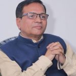 Man wearing glasses and a blue vest over a beige kurta, seated with hands clasped together in a thoughtful pose.