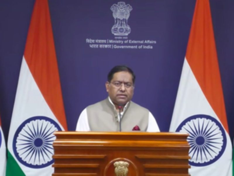 Indian government spokesperson at a wooden podium with the Emblem of India, flanked by two Indian flags behind him.