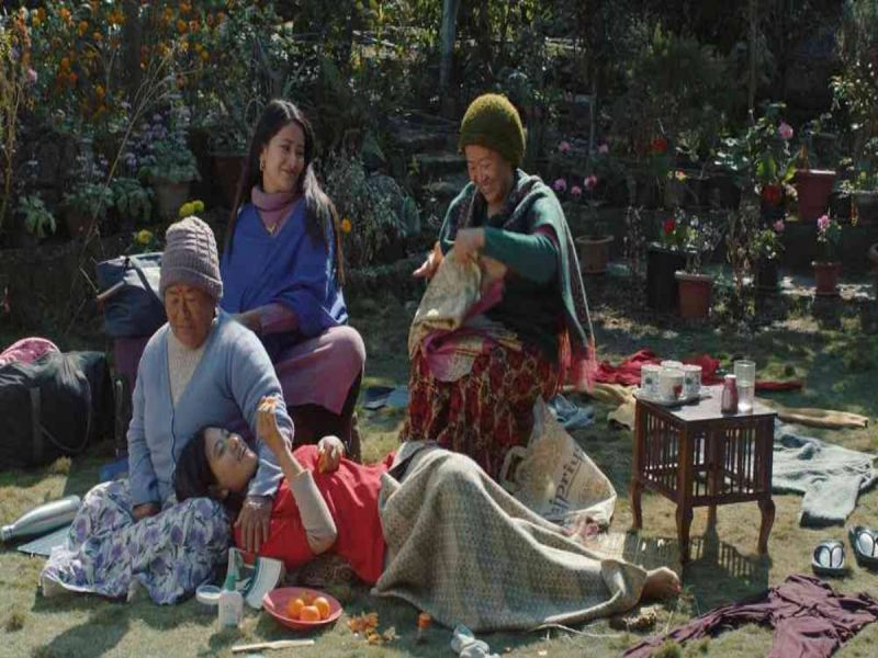 Family in a sunny garden care for an elderly person lying on a blanket while others sit nearby and share food; flowers in the background.