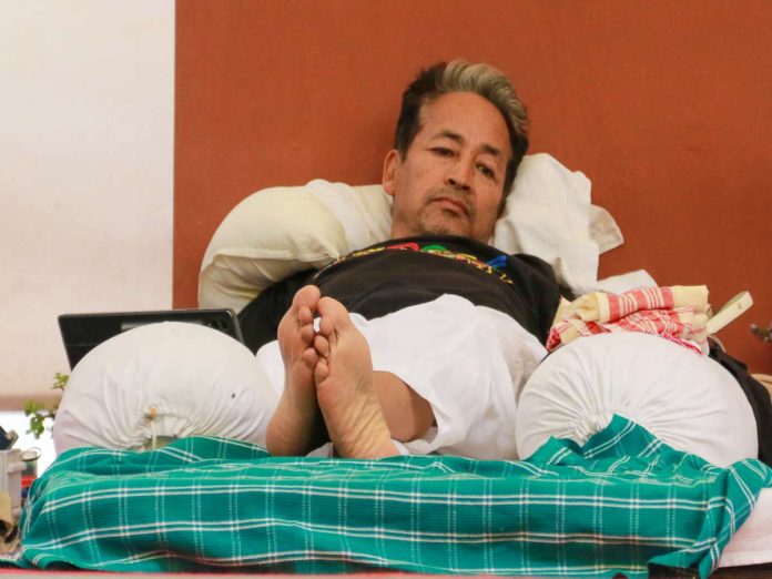 Man with gray-streaked hair lying on a bed, feet up, resting on white pillows with a green plaid blanket in the foreground.