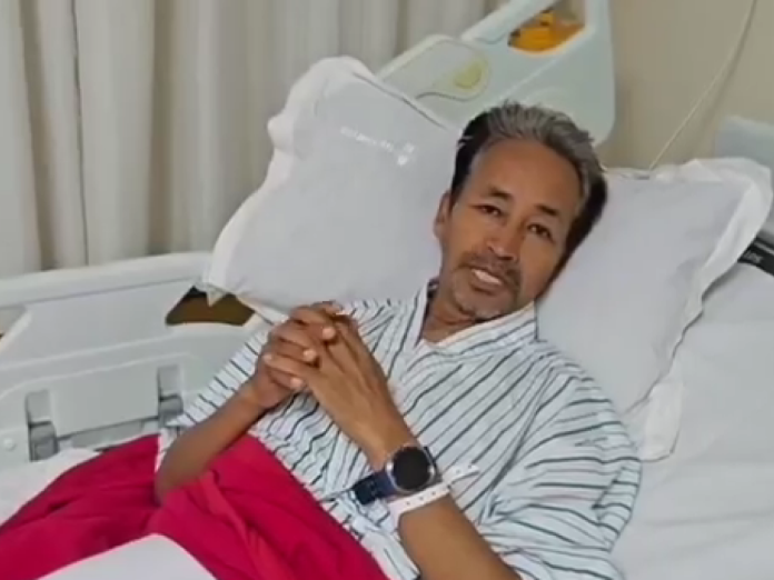 Man resting in a hospital bed, hands clasped, wearing a striped shirt and a smartwatch, with medical equipment in the background.
