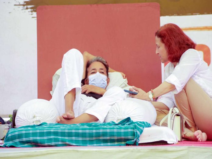 Man lying on cushions wearing a face mask while a woman with red hair shows him a smartphone nearby outside the scene's backdrop.