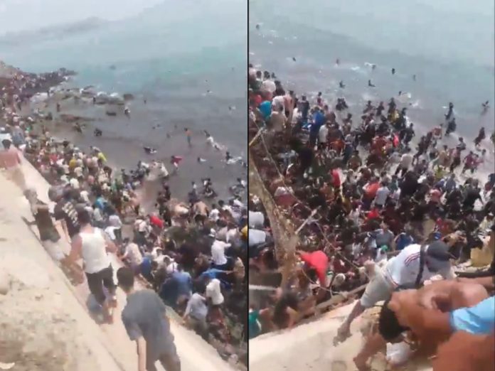 Large crowd lined up along a concrete sea wall at the shore, with people standing in the water and on the steps by the beach.