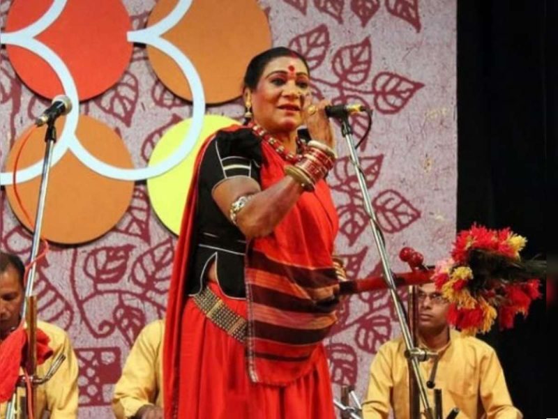teejan - bolebharat Female Indian classical singer in a red sari and gold jewelry sings into a microphone on a decorated stage, with two seated musicians in yellow shirts behind her.