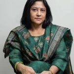 Woman in a green patterned saree sits with hands folded in her lap, looking at the camera with a calm expression.