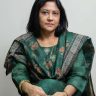 Woman in a green patterned saree sits with hands folded in her lap, looking at the camera with a calm expression.