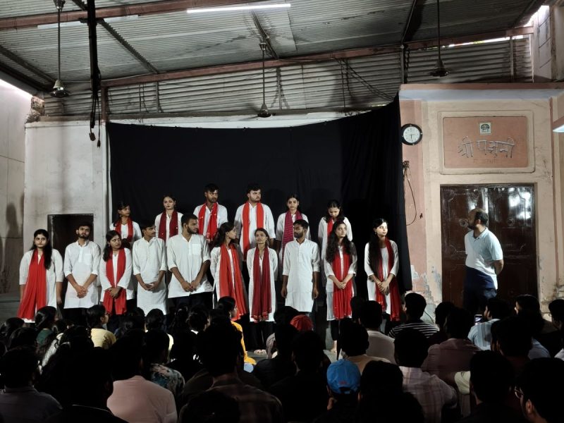 Drishaym 2 - bolebharat Group of students on a stage wearing white outfits with red scarves, performing for an audience in a hall with a black backdrop.