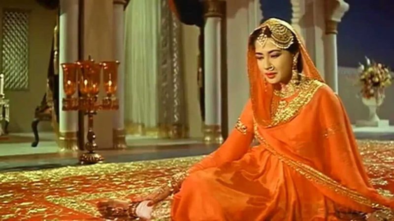 Woman in an orange traditional Indian outfit with heavy gold jewelry sits on a richly patterned red carpet in a grand palace-like setting.