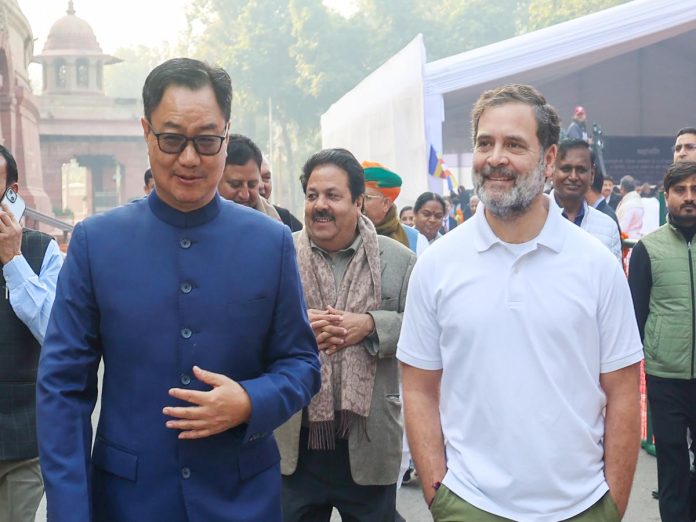 Two men walk together at an outdoor public event; one in a blue suit and glasses, the other in a white polo shirt with a beard visible behind him.