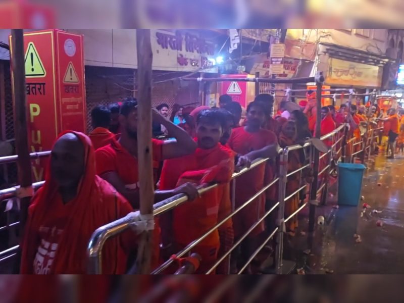 Crowd of devotees in orange robes lined up behind metal barricades on a wet street at night, waiting for a religious ceremony roughly photographed.