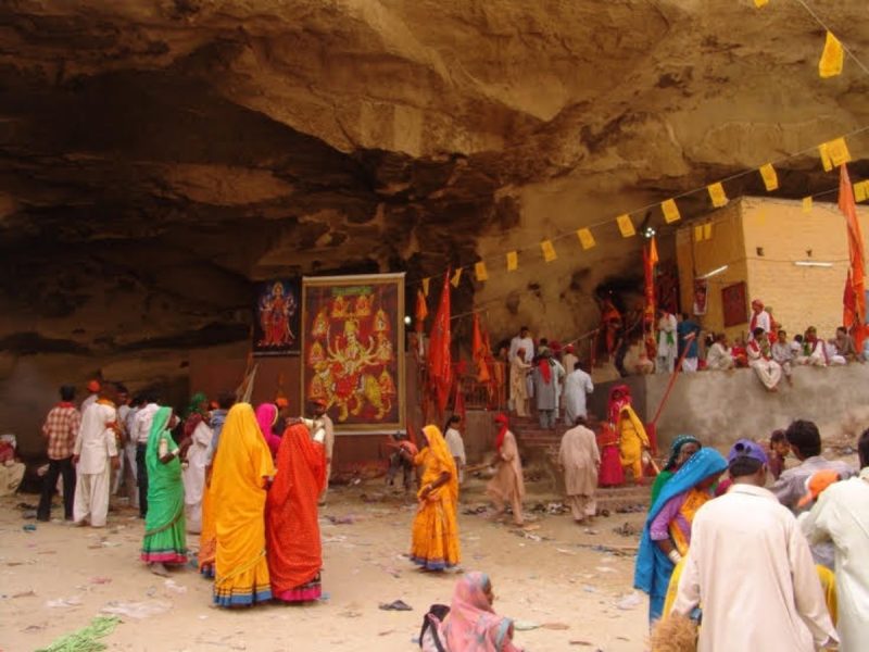Crowd of people in colorful sarees and robes gathered inside a rocky cave shrine with temple posters and yellow prayer flags outside.