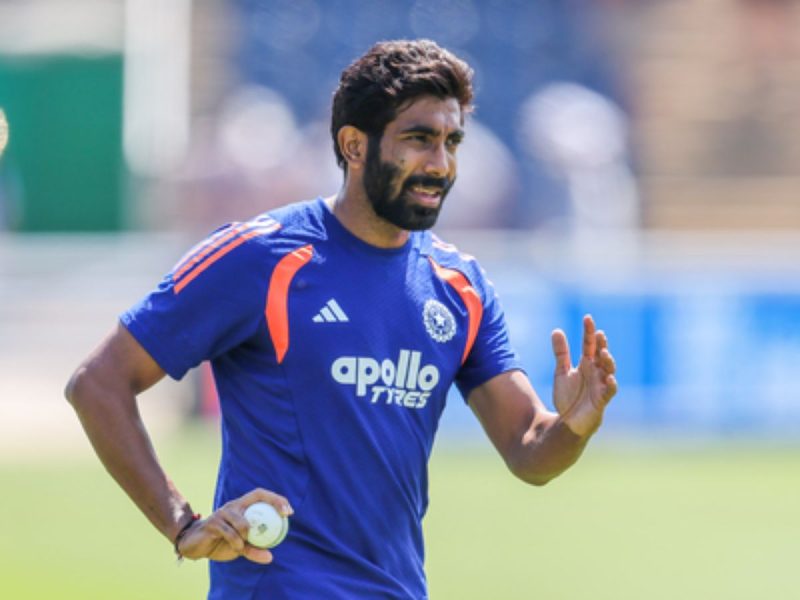Male cricketer in a blue training jersey with orange accents, holding a ball in his right hand on a practice field.