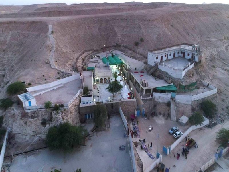 Overhead view of a cliffside complex with white buildings around a central courtyard, stairways, and a small group of people at the lower plaza.
