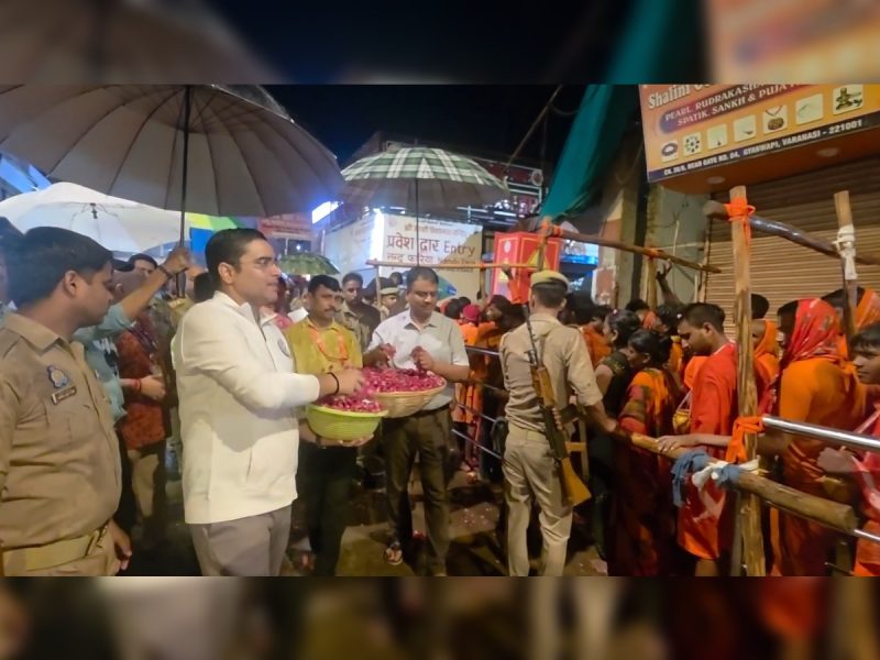 Night street scene: a man in white distributes flower petals from baskets to a line of orange-clad devotees, with police nearby under umbrellas.