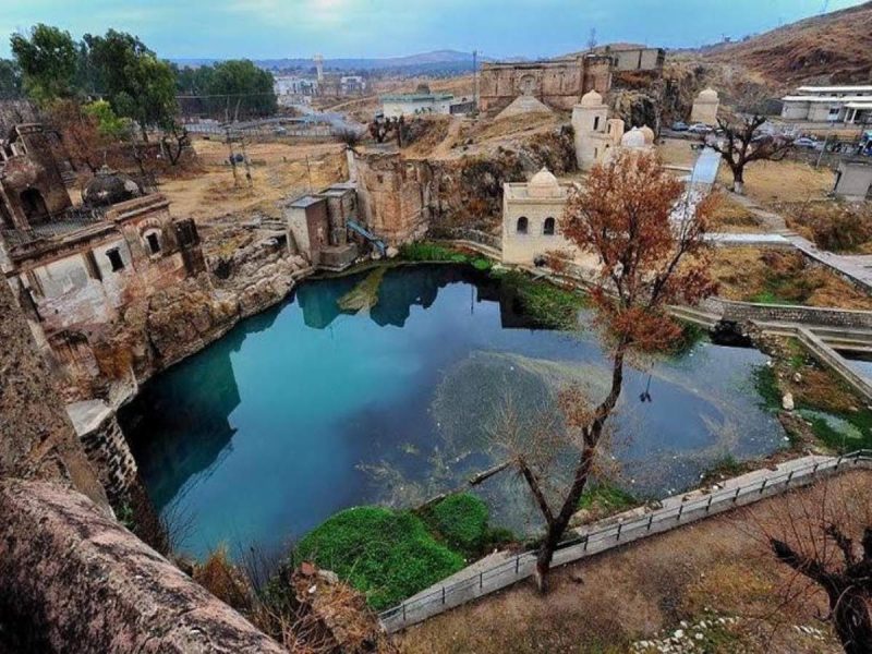 Ancient ruin complex surrounding a calm turquoise pond, with bare trees and hills in the background.