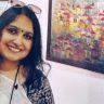 Smiling woman with a bindi, wearing a white top and patterned jacket, sunglasses on head, indoors beside a colorful painting of houses