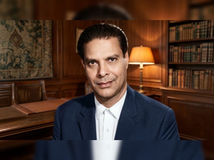 Man in a navy suit and white shirt in a wood-paneled library, looking at the camera.