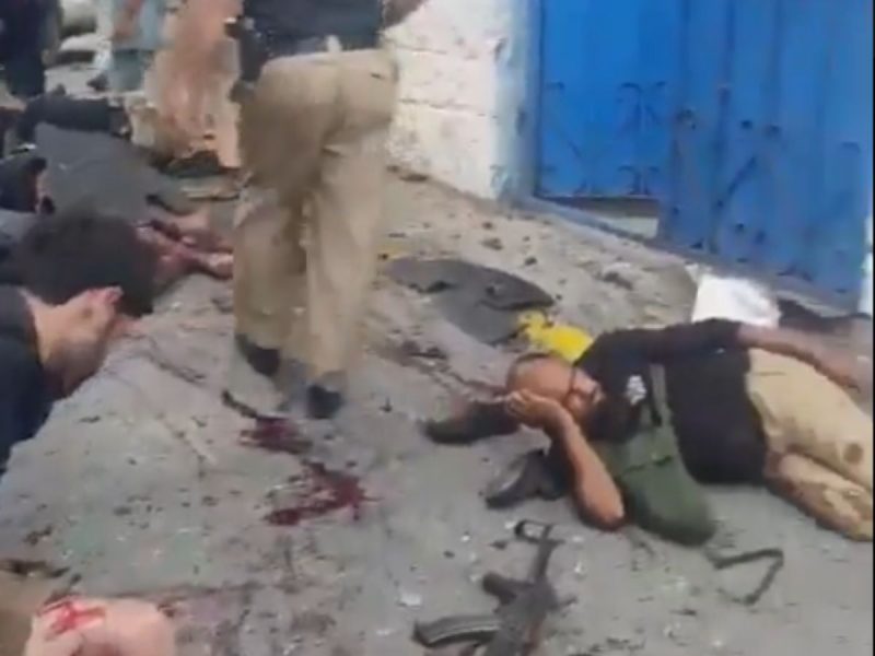 Civilian injuries on a rubble-strewn street near a blue wall; people lie on the ground while others stand nearby, suggesting aftermath of an explosion or collapse (non-graphic).