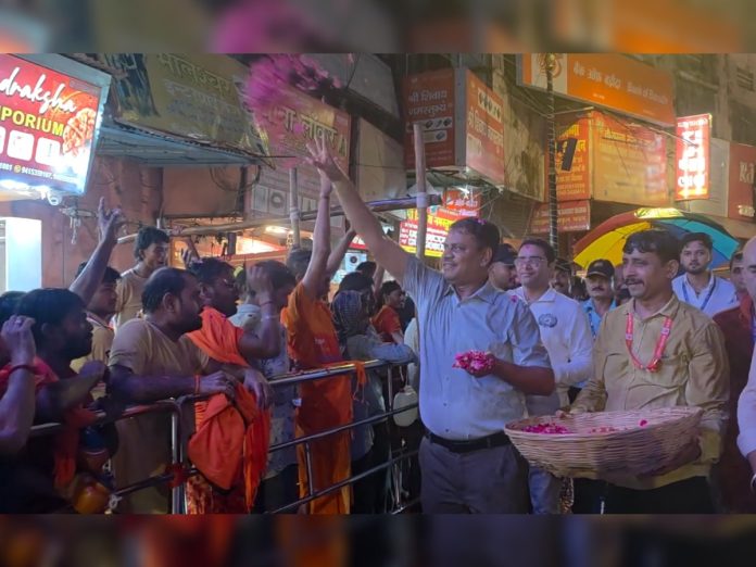 Man in a light blue shirt raises his hands toward a cheering crowd at a street festival, holding pink flower petals; monks in orange robes stand nearby.