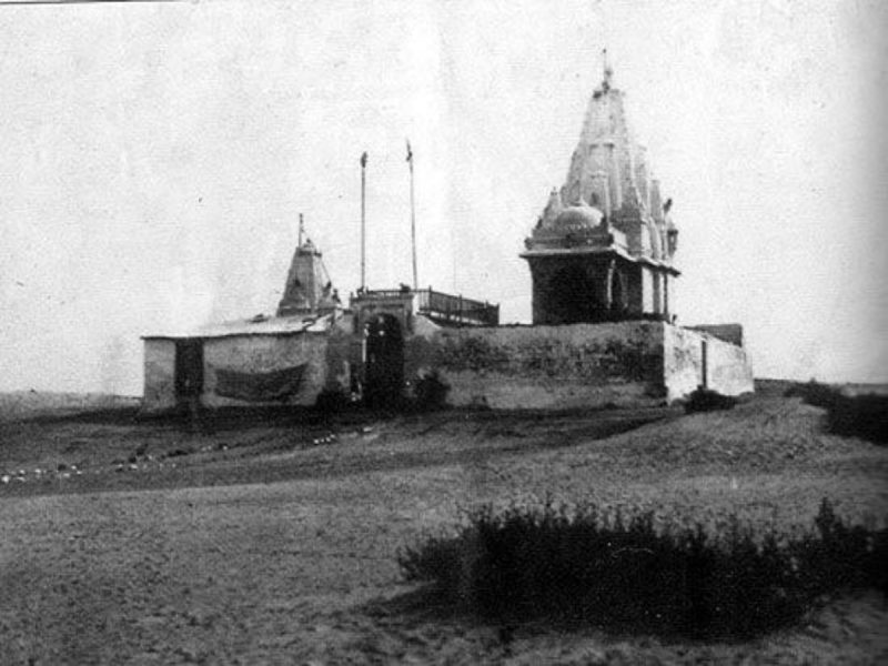 Black-and-white photo of a small temple with an ornate spire on a hill, surrounded by stone walls and a dirt path, sparse landscape.